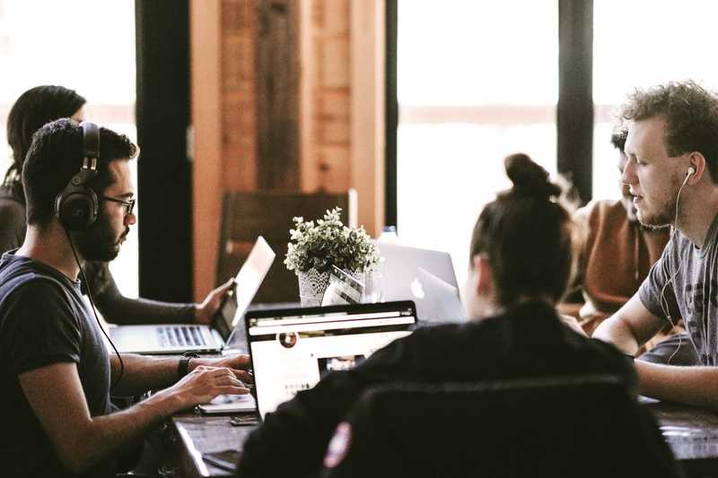 Eine Gruppe junger Menschen sitzt zusammen an einem großen Tisch. Alle arbeiten an ihren Laptops.