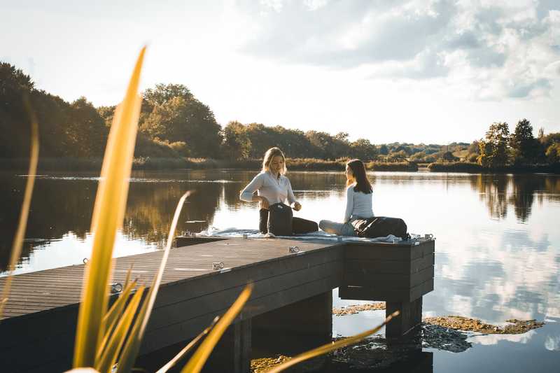 Zwei Frauen sitzen auf einem Stegan einem Teich in der untergehenden Sonne und machen ein Picknick.