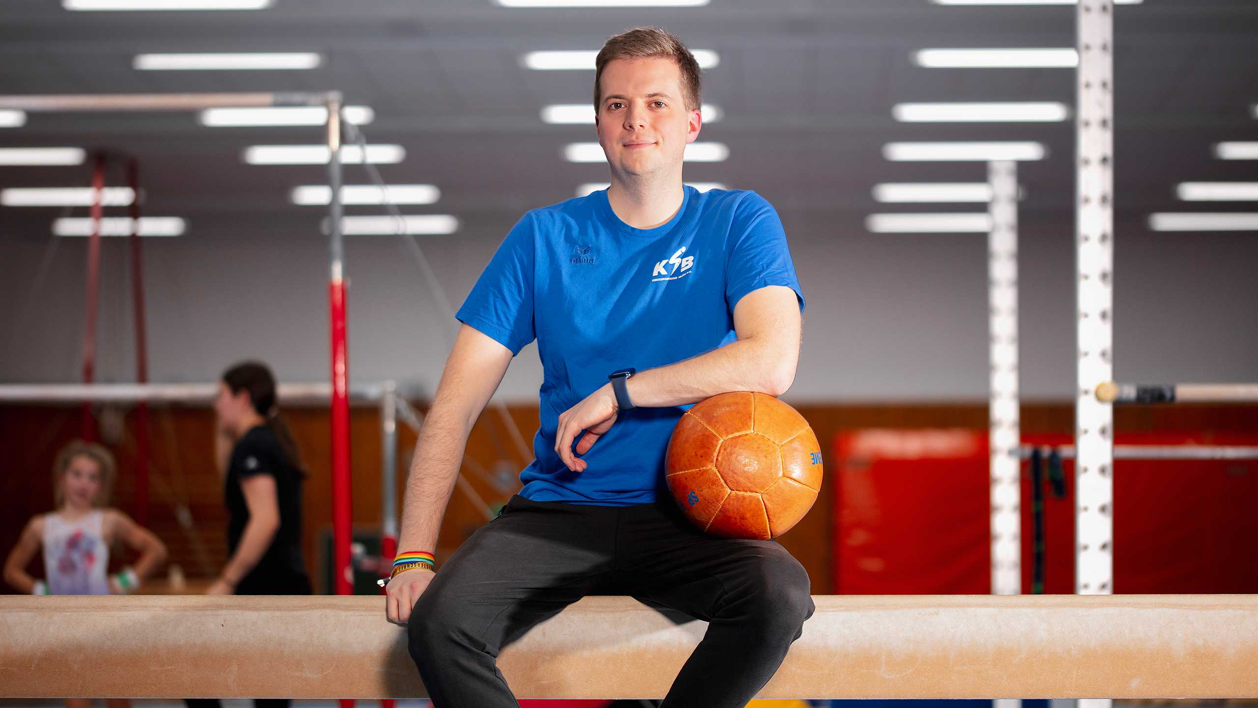 Ein junger Mann sitzt mit einem Medizinball in der Hand auf einem Schwebebalken.