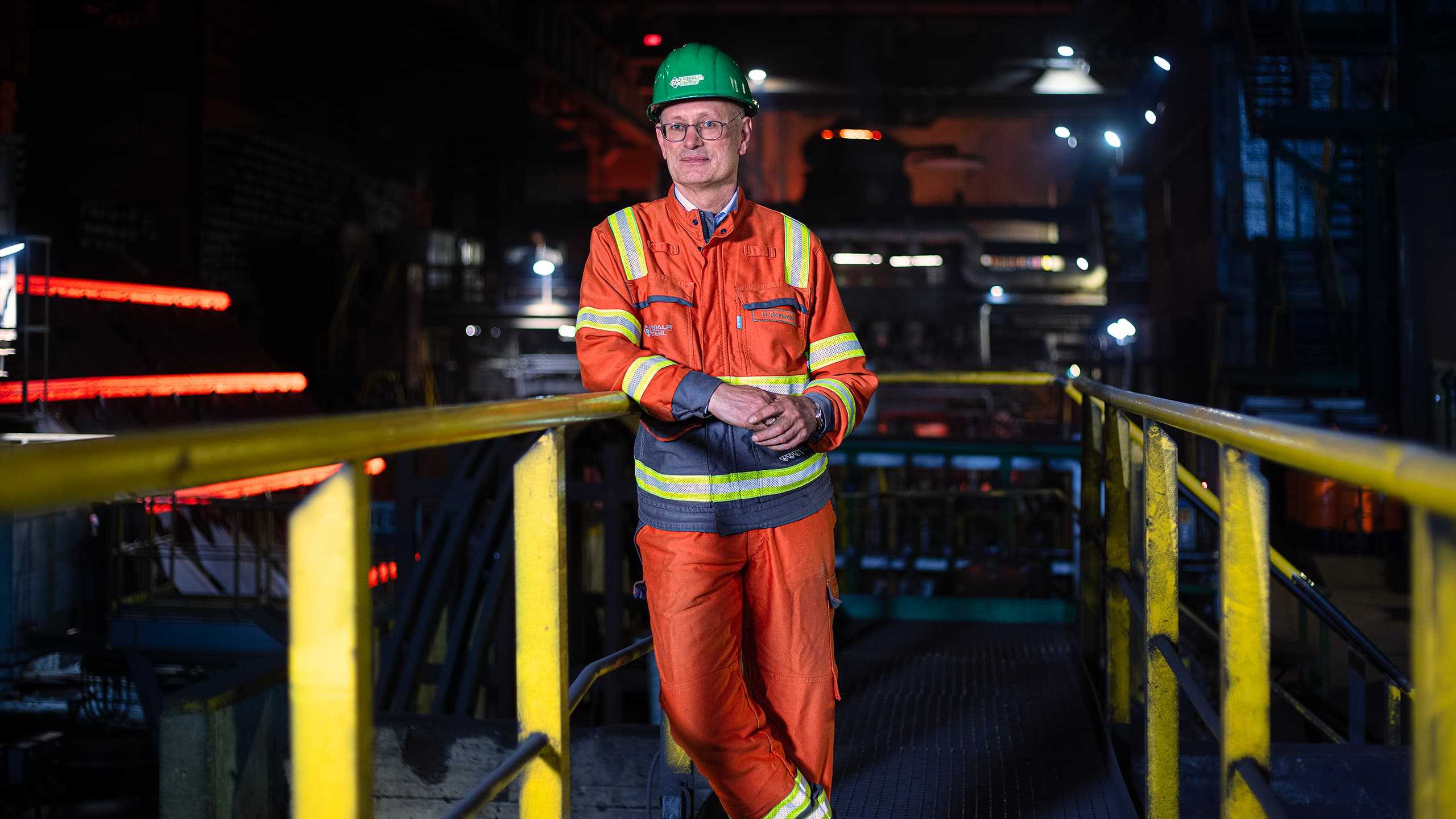 Ein Mann in einem orangefarbenen Schutzanzug und grünem Helm steht in einer Stahlfabrik. Er lehnt an einem Geländer, umgeben von industrieller Ausrüstung und rot glühendem Stahl im Hintergrund.