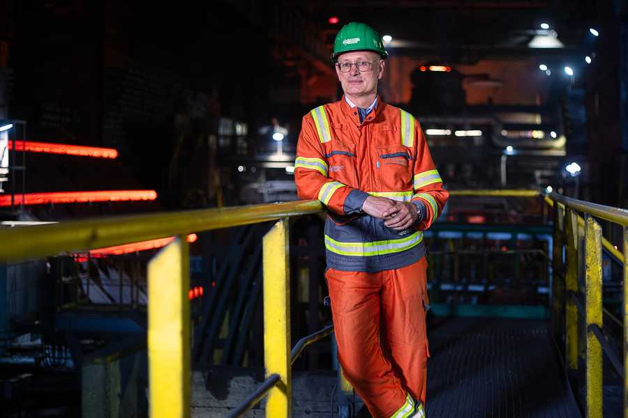 Ein Mann in einem orangefarbenen Schutzanzug und grünem Helm steht in einer Stahlfabrik. Er lehnt an einem Geländer, umgeben von industrieller Ausrüstung und rot glühendem Stahl im Hintergrund.