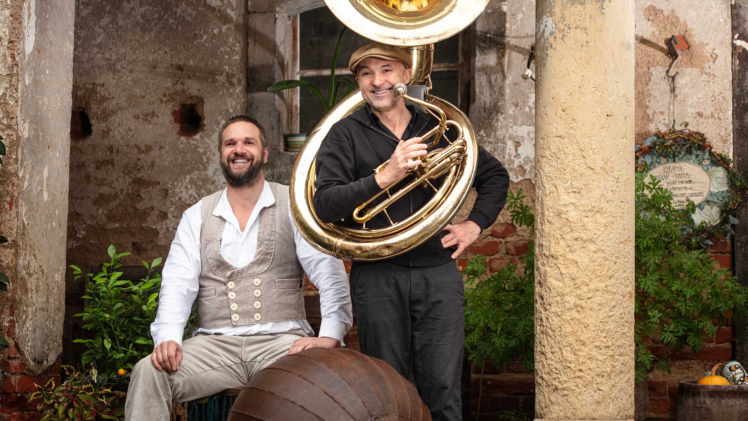 Ein Mann mit Steinmetzkluft und ein Musiker mit einer großen Tuba sitzen vor einem alten Gebäude mit Säulen. Beide lachen fröhlich.