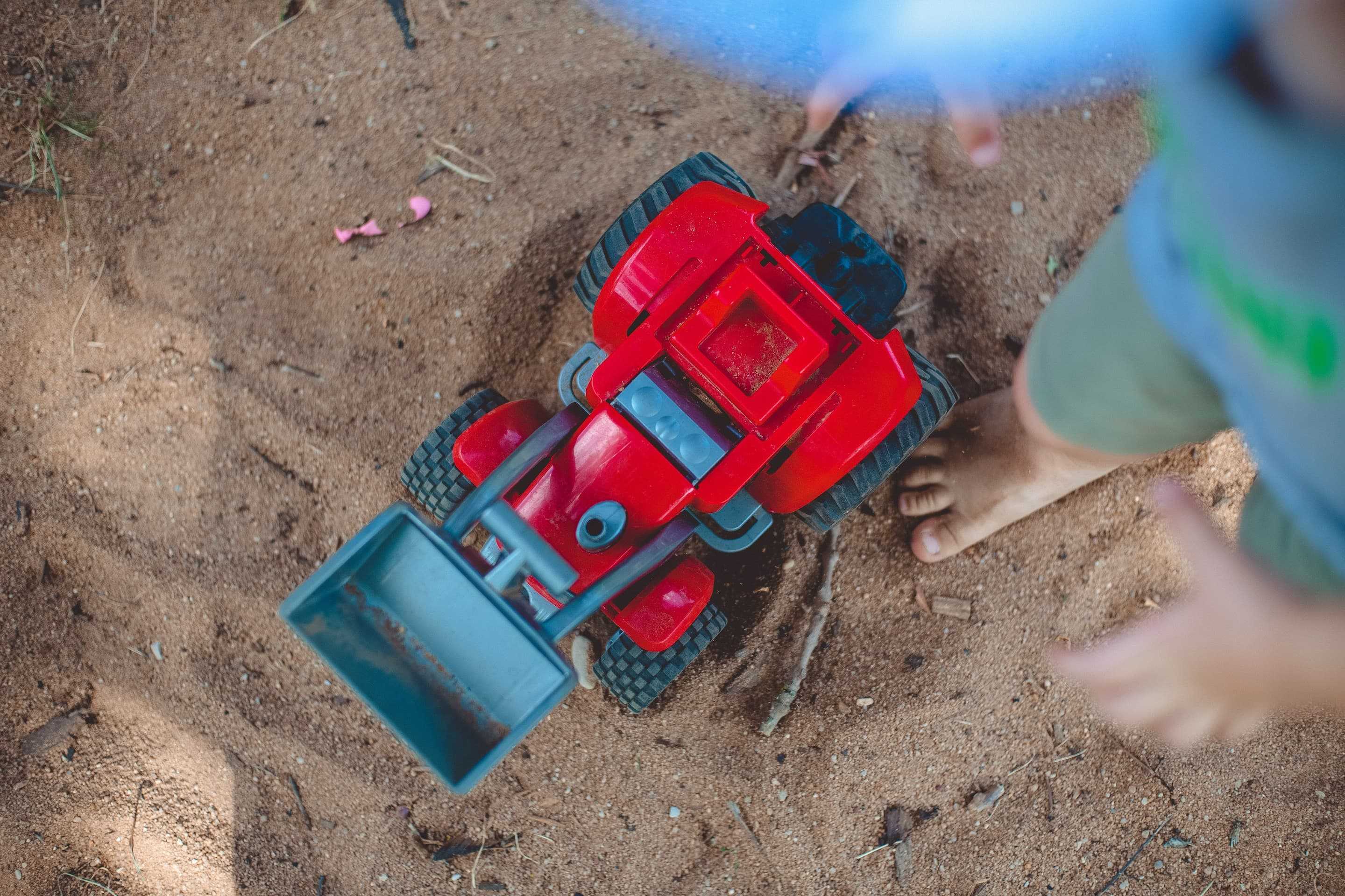 Ein roter Spielzuegbagger in einem Sandkasten von oben fotografiert. Daneben steht ein Kind, es ist barfuss und man kann nur seine Füße sehen.